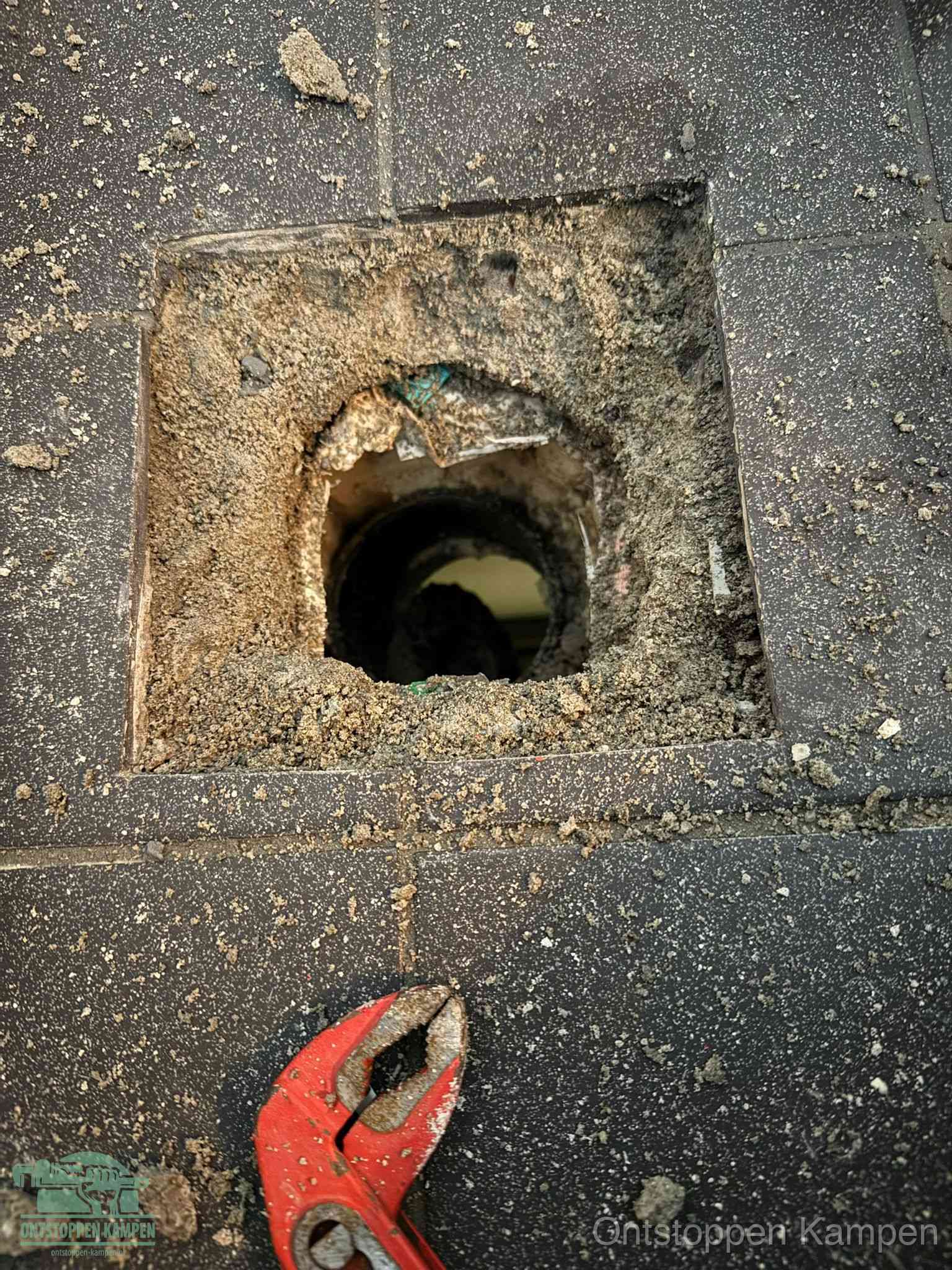 Een geopend rioolputje in een betontegelvloer met losgemaakt rooster en een zichtbare ronde afvoerbuis, het achterhalen waar de afvoerverstopping zit in Kampen