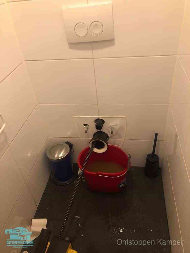 Een ontstopte wand-wc met verwijderde toiletpot waar een ontstoppingsveer in de wanduitgang verdwijnt, het achterhalen waar de verstopping zit in Kampen
