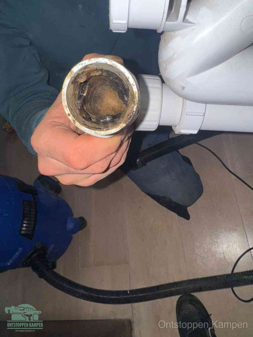 Een hand houdt een losgekoppeld PVC-afvoerbuisstuk vast dat van binnen volledig is dichtgeslibd met vet, vuil en aanslag, de meest voorkomende oorzaak van een verstopte afvoer in Kampen