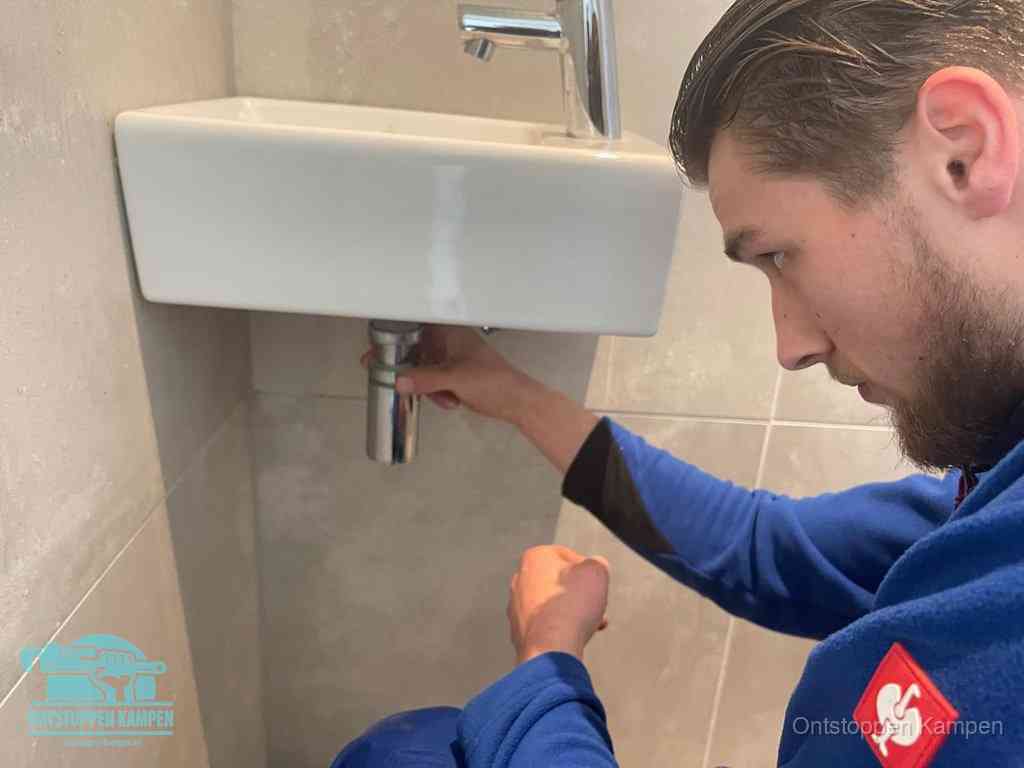 Een monteur draait met de hand de zwanenhalssifon onder een fonteinwasbak in de toiletruimte los om de bron van rioollucht in Kampen te vinden