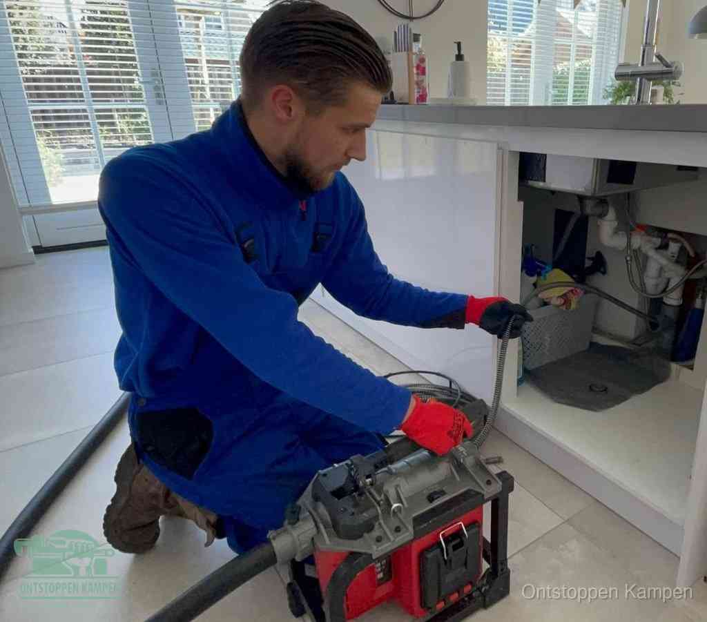 Een monteur met rode handschoenen knielt in een keuken en voert een elektrische ontstoppingsveer in tijdens het professioneel afvoer ontstoppen in Kampen
